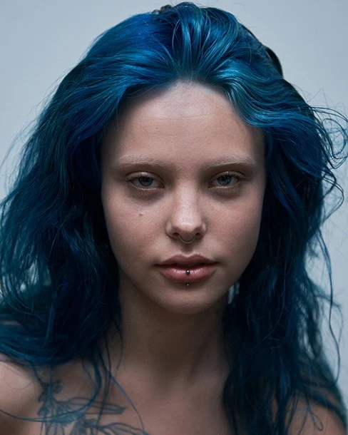 Striking portrait of a young adult woman with vibrant blue hair, fair skin, and a tattoo on her chest, set against a neutral background.