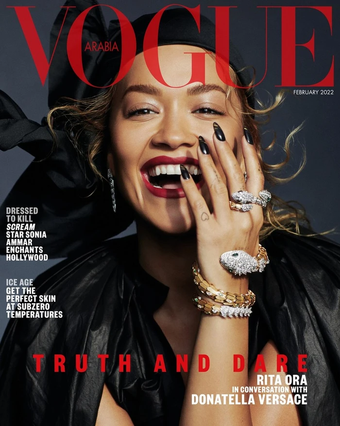 Rita Ora laughing with a hand near her face, wearing sparkling diamond snake rings, a black outfit, and red lipstick.