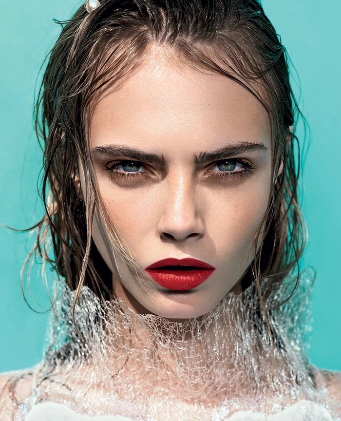 Close-up portrait of a woman with wet hair, bold red lipstick, and a textured white collar against a turquoise background.