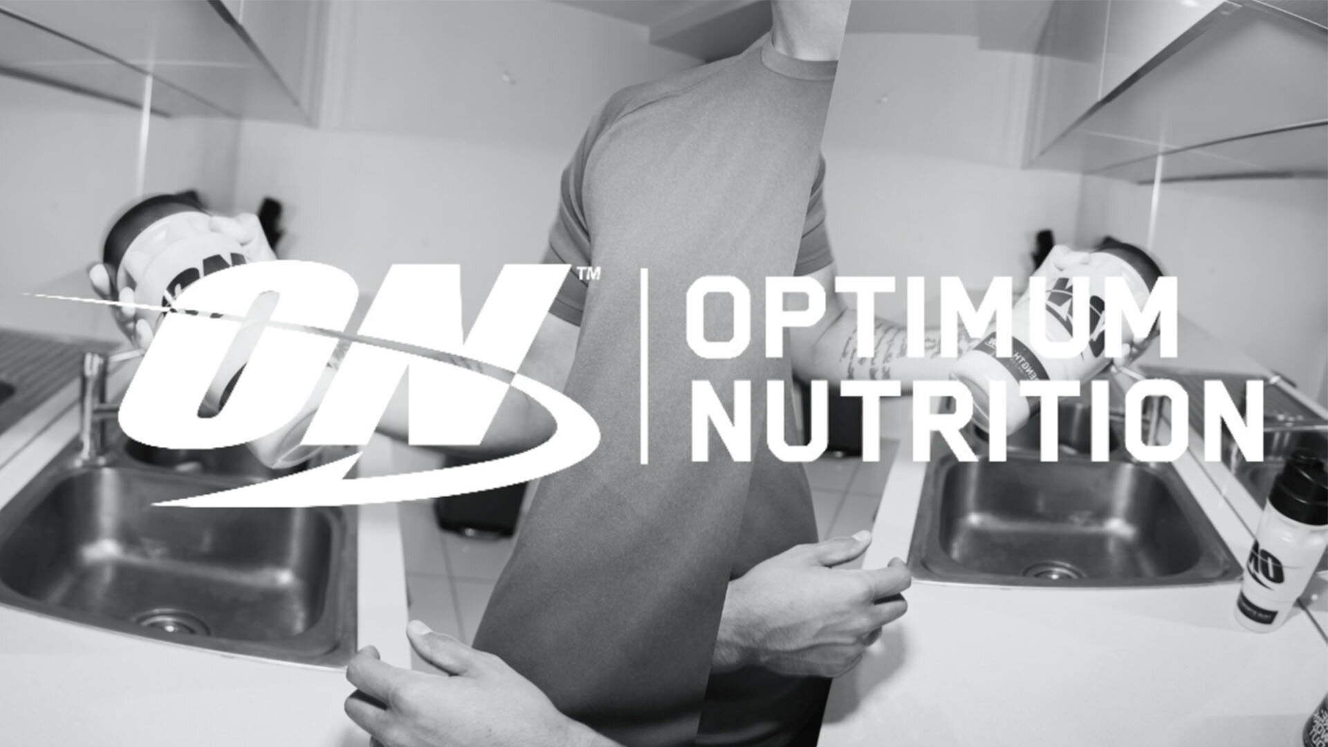 A man in front of a sink holds two containers of Optimum Nutrition supplements in a black and white, eye-level shot.