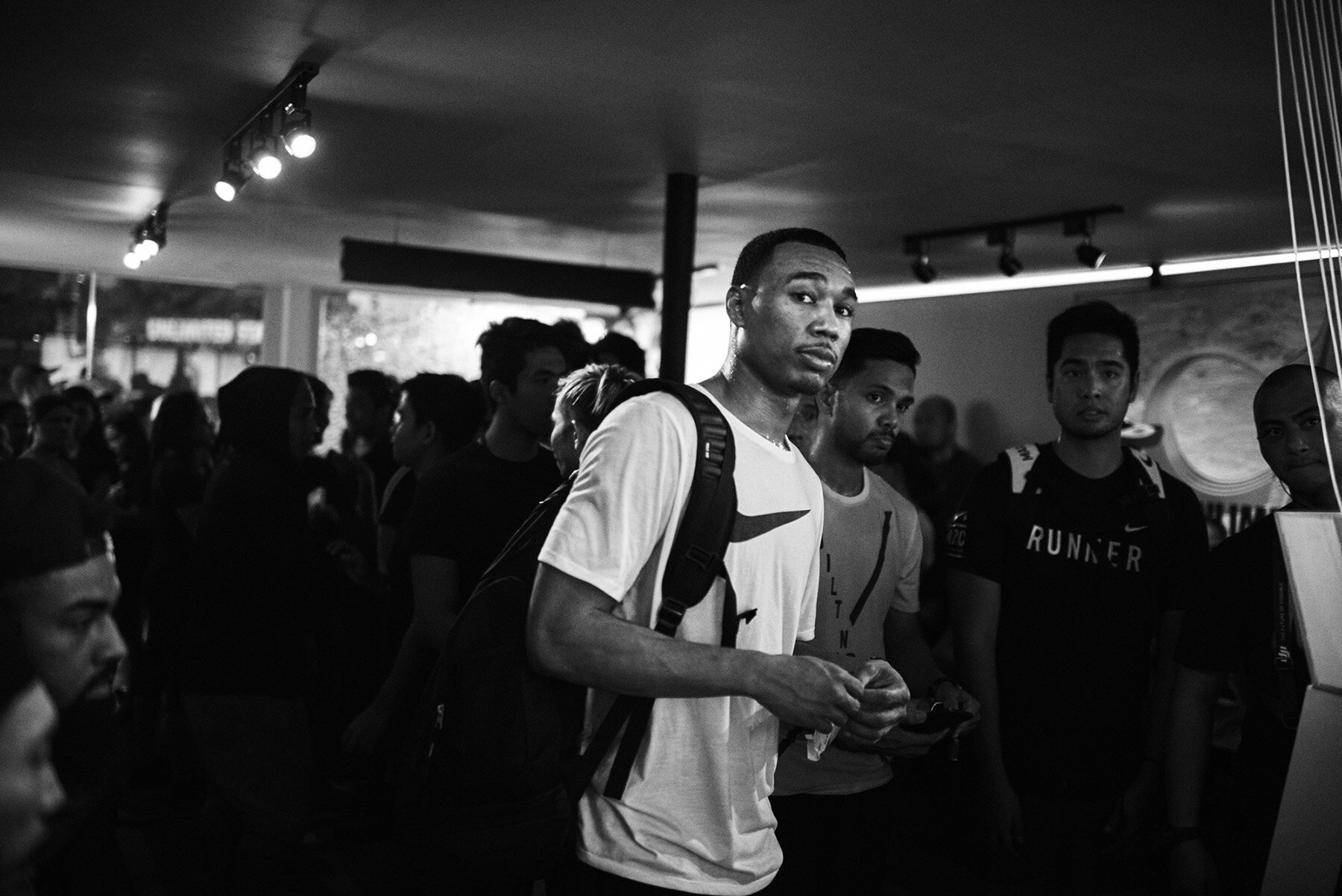 Indoor crowd scene in black and white features a male in white t-shirt with a backpack looking forward, surrounded by other people.