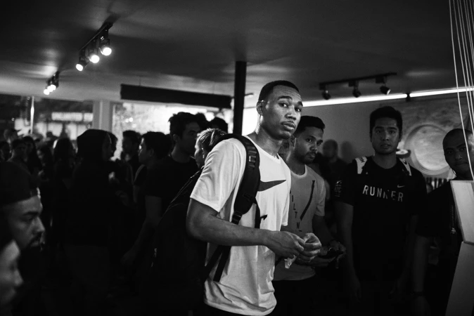Indoor crowd scene in black and white features a male in white t-shirt with a backpack looking forward, surrounded by other people.