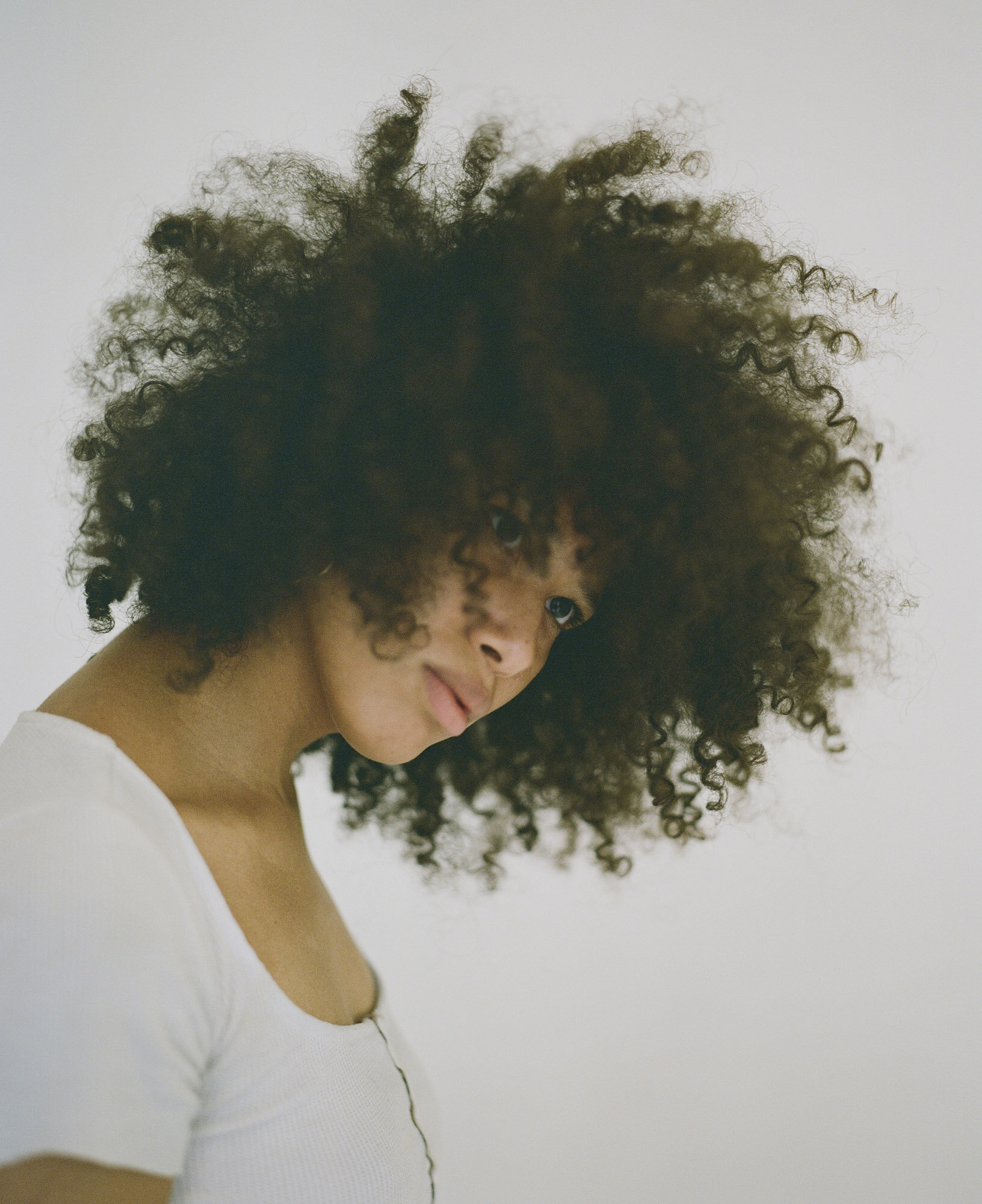 Portrait of a young adult woman with curly afro hair and a white top, posing against a plain, light background in a studio setting.