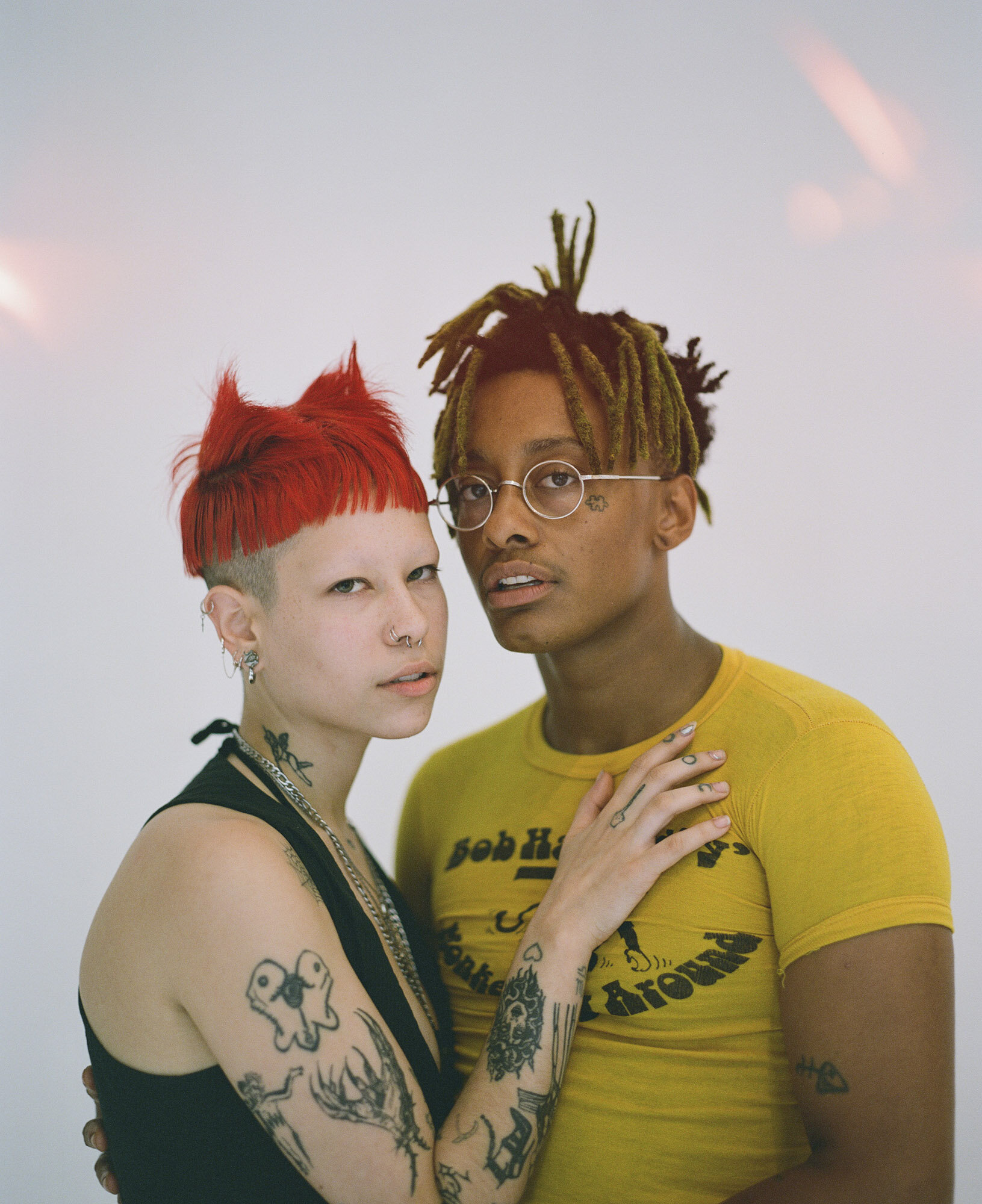 Close-up portrait of a young couple in a photo studio. The woman has short red hair and tattoos. The man has dreadlocks and round glasses.
