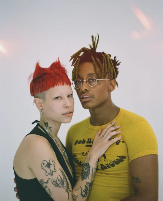 Close-up portrait of a young couple in a photo studio. The woman has short red hair and tattoos. The man has dreadlocks and round glasses.