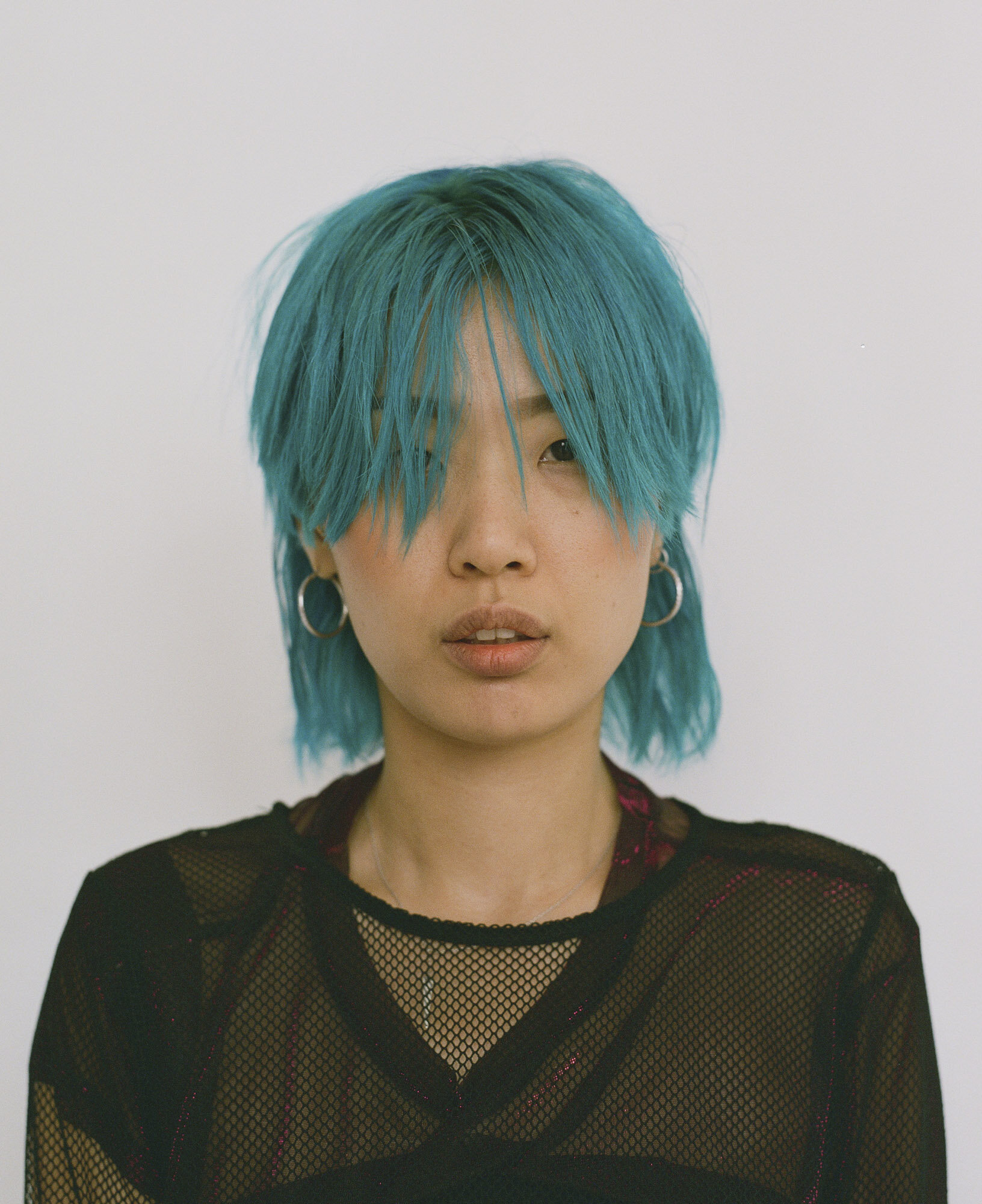 Headshot portrait of an Asian woman with vibrant teal hair, wearing a mesh top, against a plain white background. Fashionable studio shot.