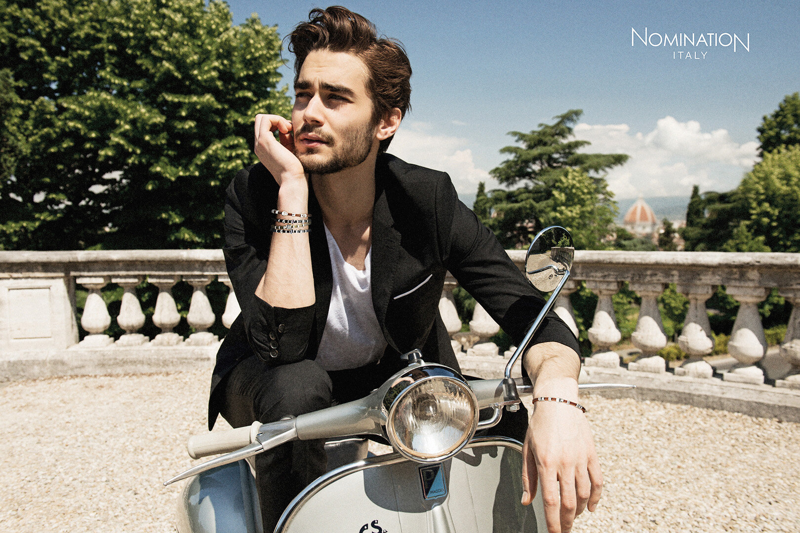 Adult male poses on a gray scooter wearing jewelry, a blazer, and white t-shirt against a cityscape background on a sunny day.