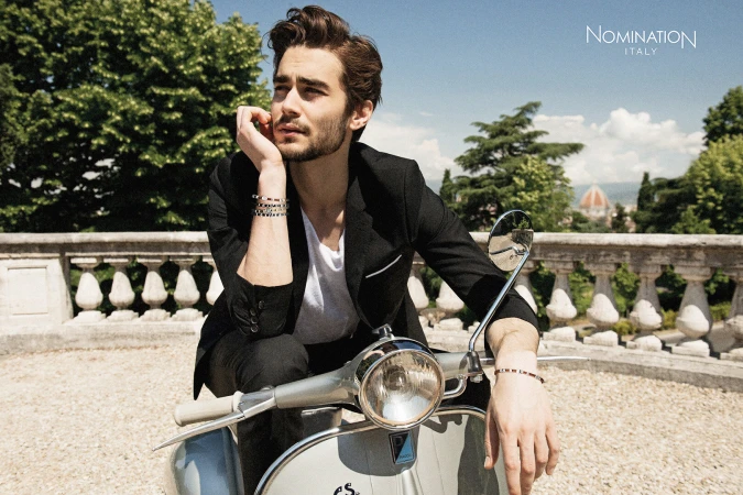 Adult male poses on a gray scooter wearing jewelry, a blazer, and white t-shirt against a cityscape background on a sunny day.