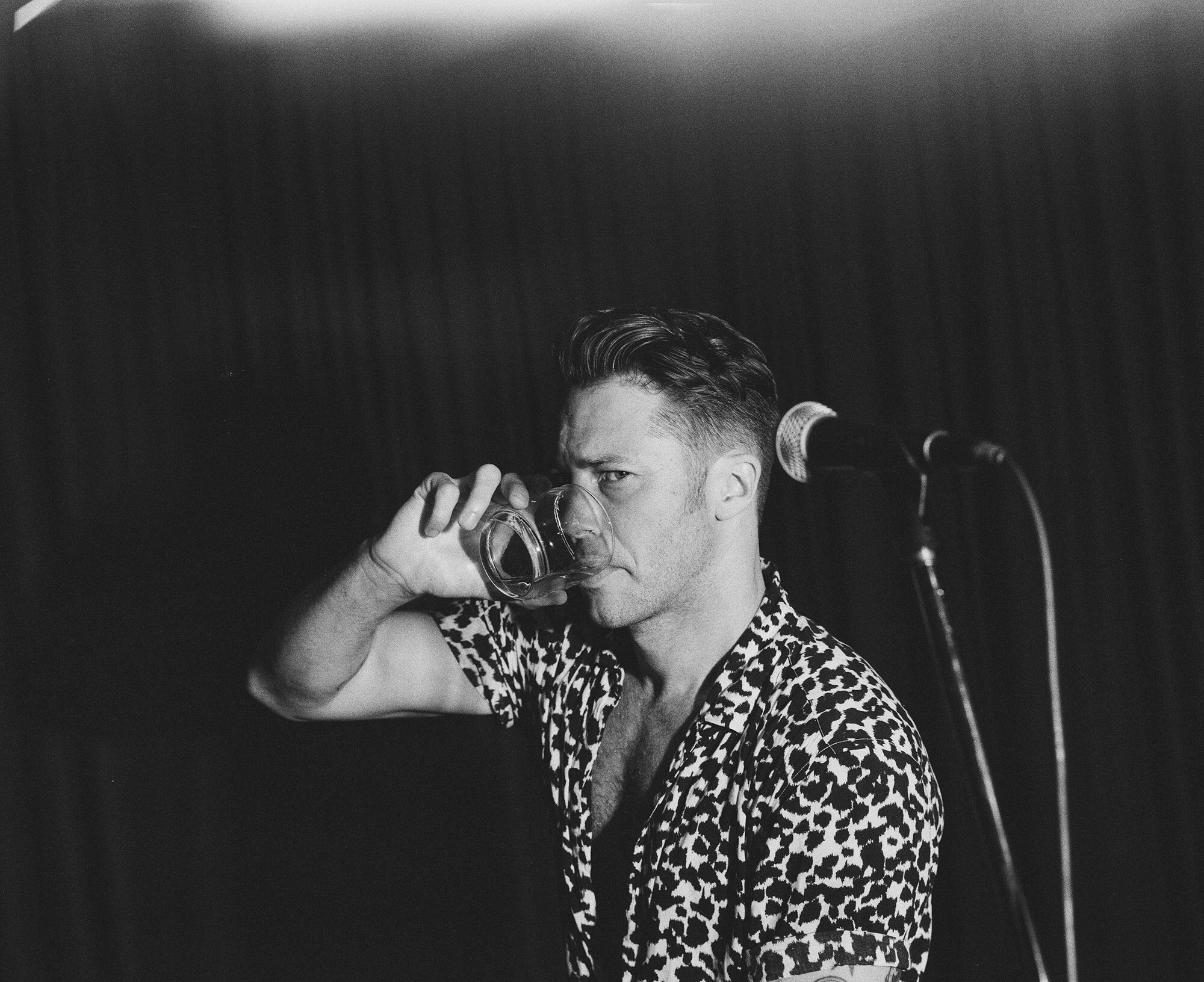 Black and white portrait of a man drinking from a glass in front of a microphone on stage, with a dark background and animal-print shirt.