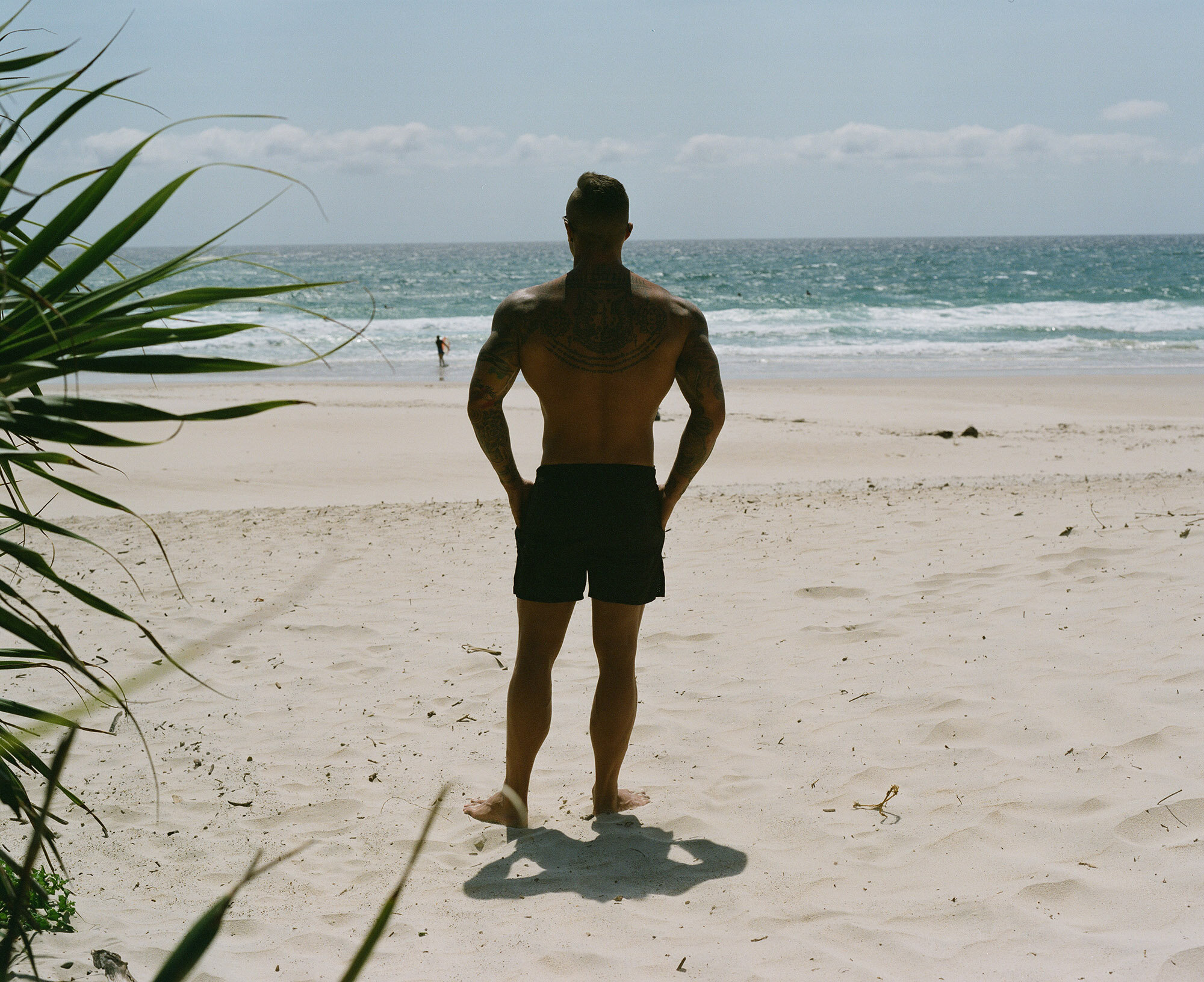 Adult male stands on a sunny beach facing the ocean, with vegetation in the foreground, captured in a landscape orientation.