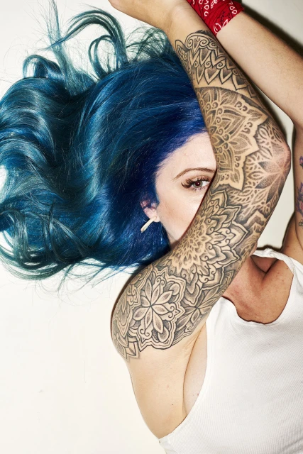 Woman with bright blue hair and arm tattoos lying on a white surface, face partially obscured by arm, wearing white tank top.