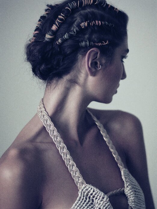Profile shot of a woman with styled dark hair featuring coils, wearing a woven top, against a neutral background in soft lighting.