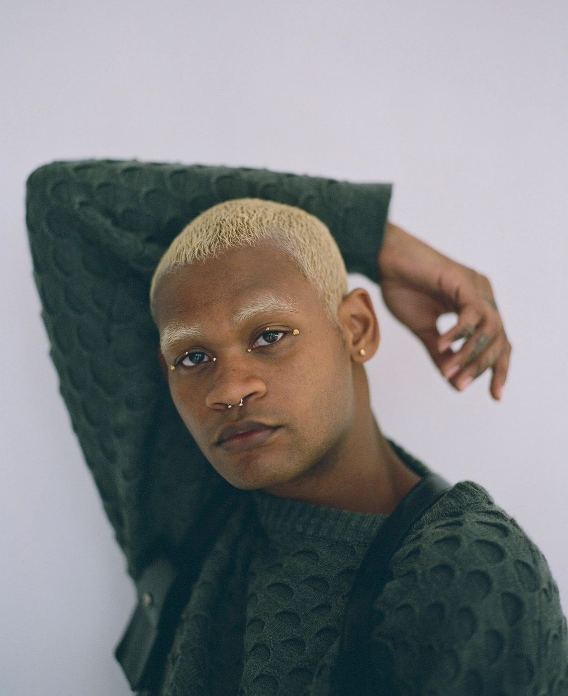 Portrait of a black man with bleached blonde buzzcut hair wearing a dark green patterned sweater against a neutral background