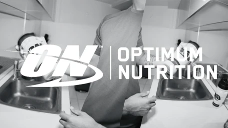 Monochrome commercial photograph featuring a person in a kitchen setting holding protein supplement tubs, photographed by Ben Cook.