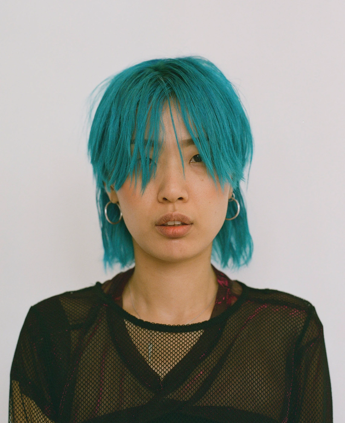 Portrait of a young person with vibrant turquoise hair and silver hoop earrings wearing a black mesh top against a white background.