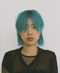 Portrait of a young person with vibrant turquoise hair and silver hoop earrings wearing a black mesh top against a white background.