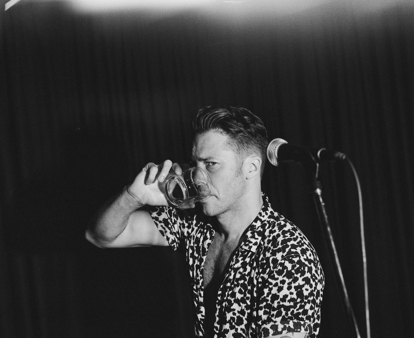 Monochrome portrait of a man in a patterned short-sleeve shirt drinking from a glass, with a microphone in the background.