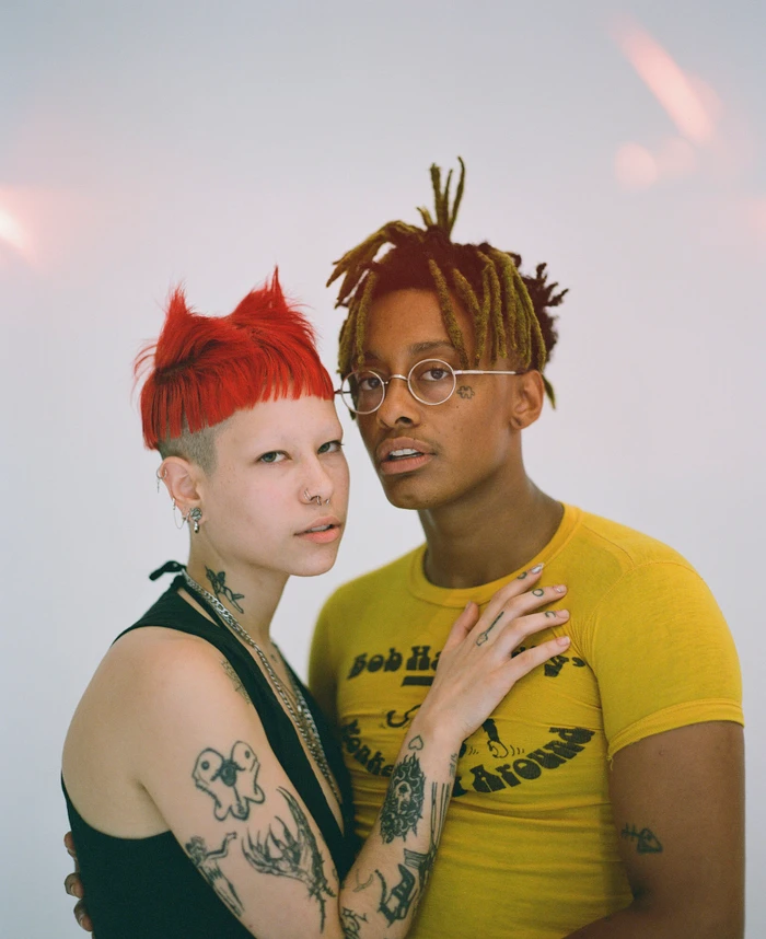 Portrait of two people with tattoos, one wearing a yellow shirt and glasses, the other with bright red hair and a black top.