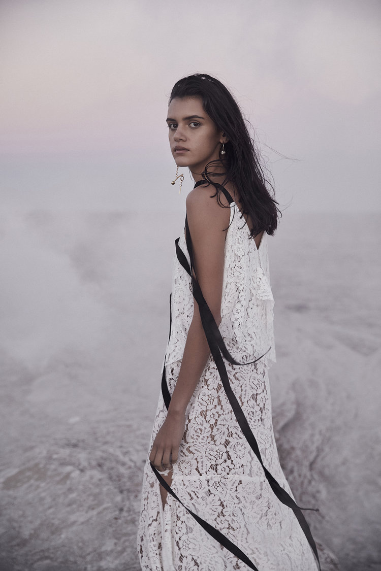 A young female model poses in a white lace dress with black ribbon against a neutral outdoor backdrop, showcasing fashion and style.