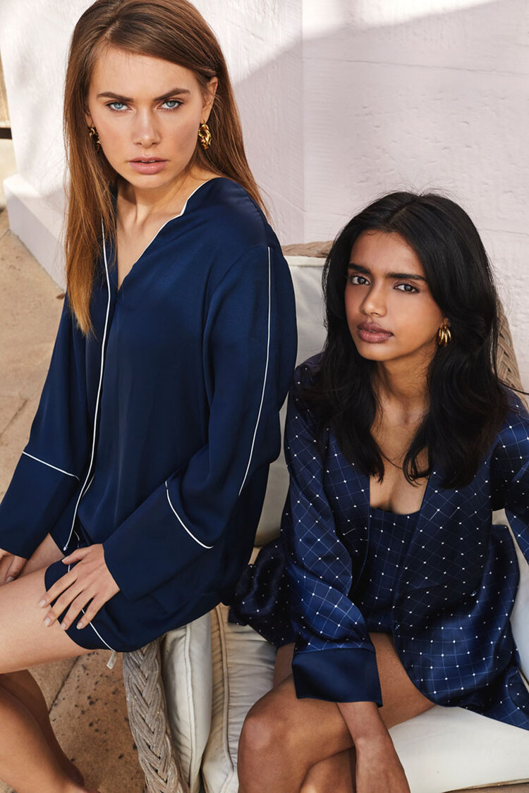 Two adult female models pose indoors, wearing blue patterned pajamas. One woman wears a long-sleeved top; both sit on a couch in a relaxed style.