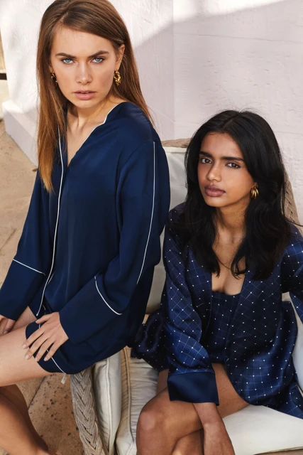 Two adult female models pose indoors, wearing blue patterned pajamas. One woman wears a long-sleeved top; both sit on a couch in a relaxed style.