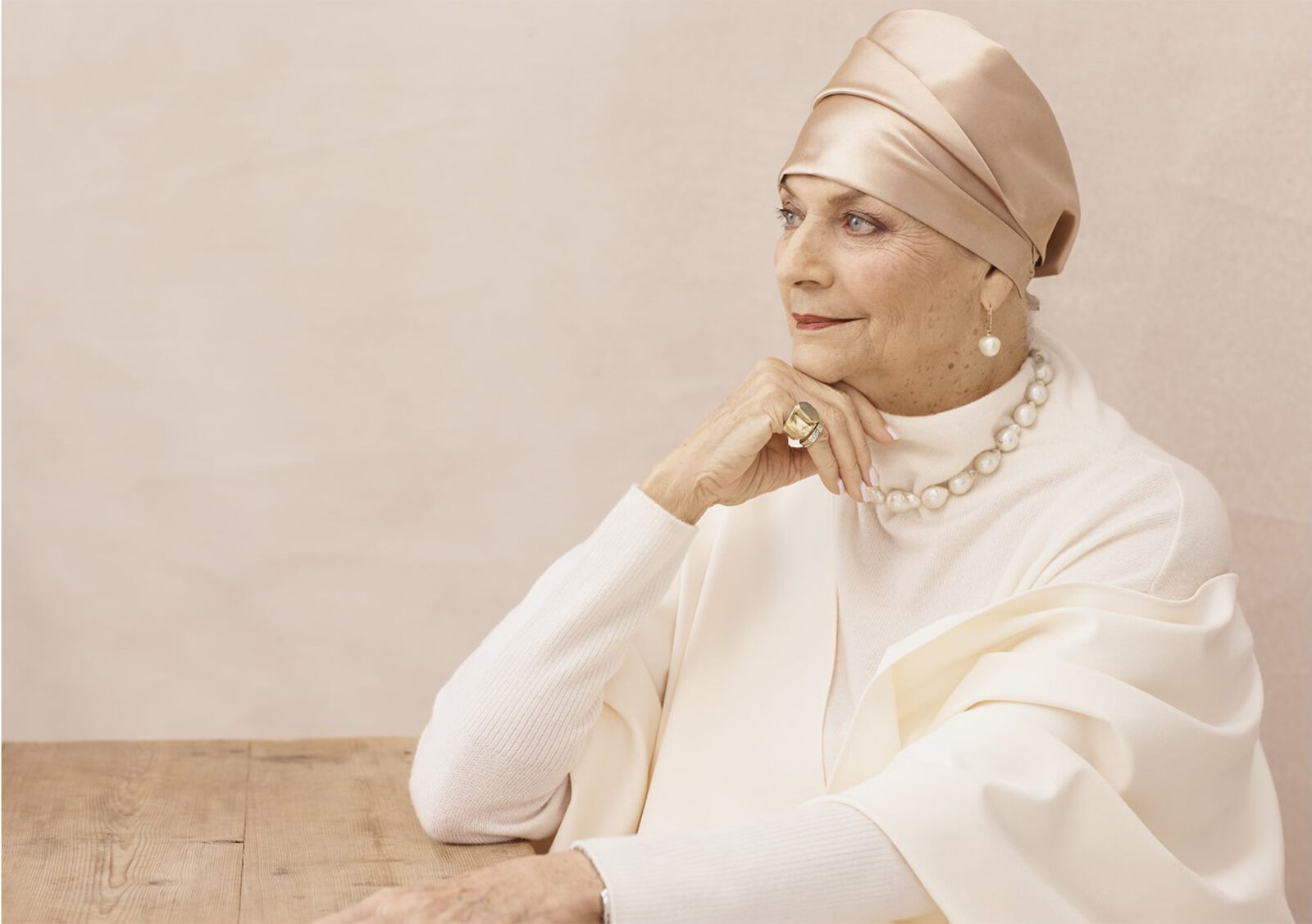 Elegant elderly woman in a headscarf wearing pearl jewelry, seated at a wood table against a neutral backdrop in a sophisticated portrait.