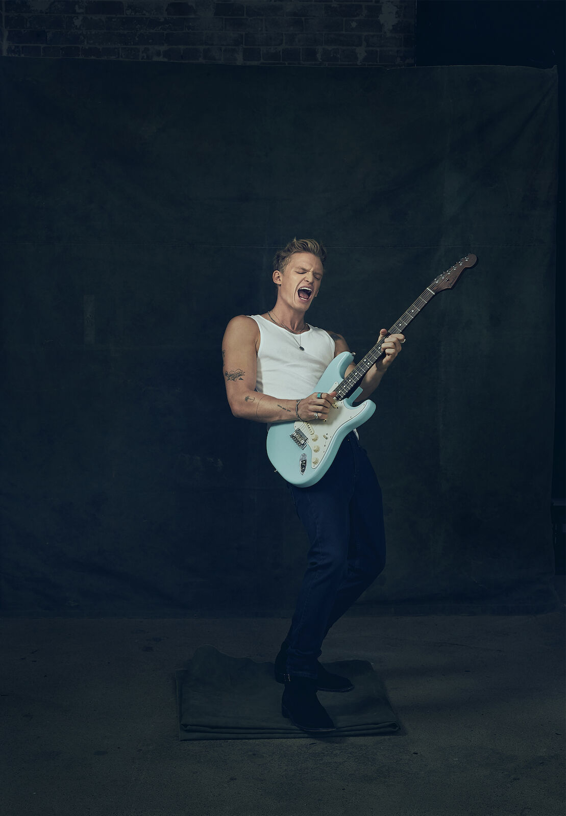 A man passionately plays an electric guitar in a studio setting. He wears a white tank top and dark pants while standing on a gray mat.