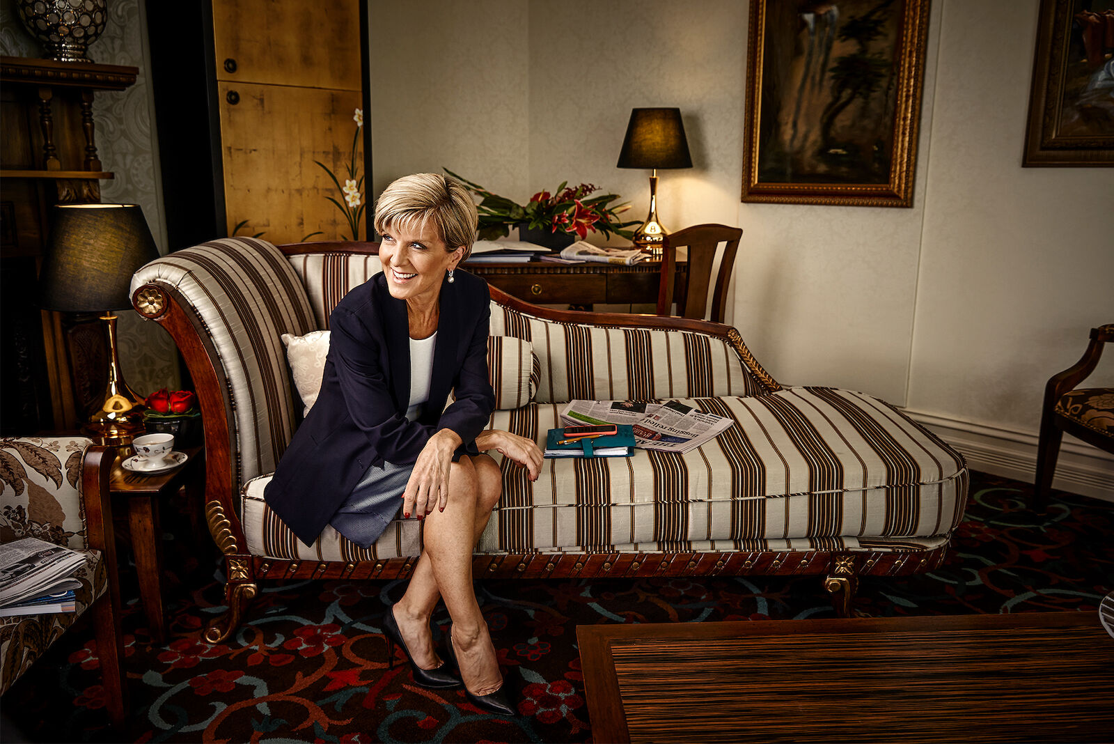 Adult female in a blazer and skirt sits crossed-legged on a striped lounge with documents in an ornate room with a patterned rug.