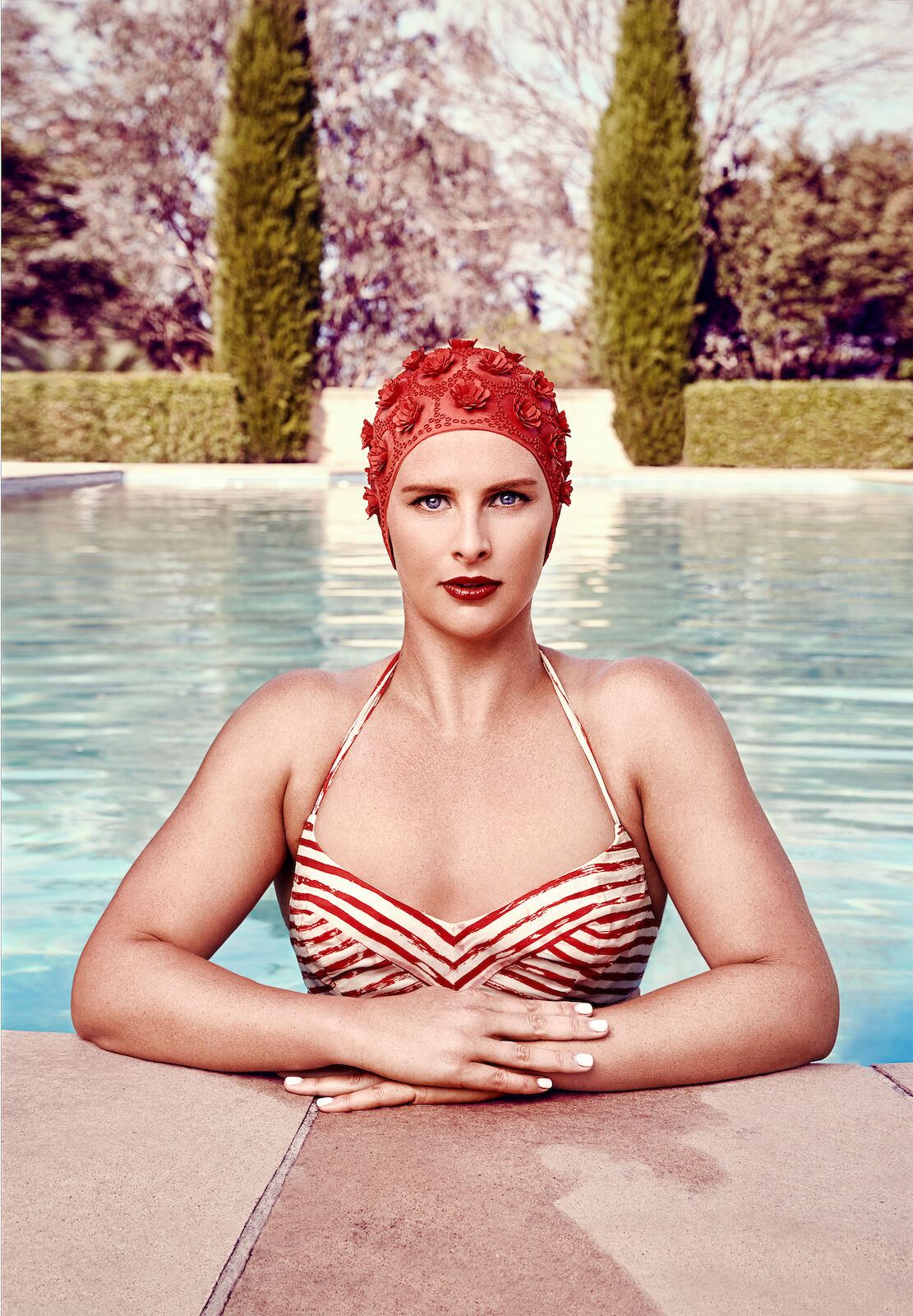 Woman in a striped swimsuit and red swim cap posing in a pool with trees and green hedges in the background on a sunny day.