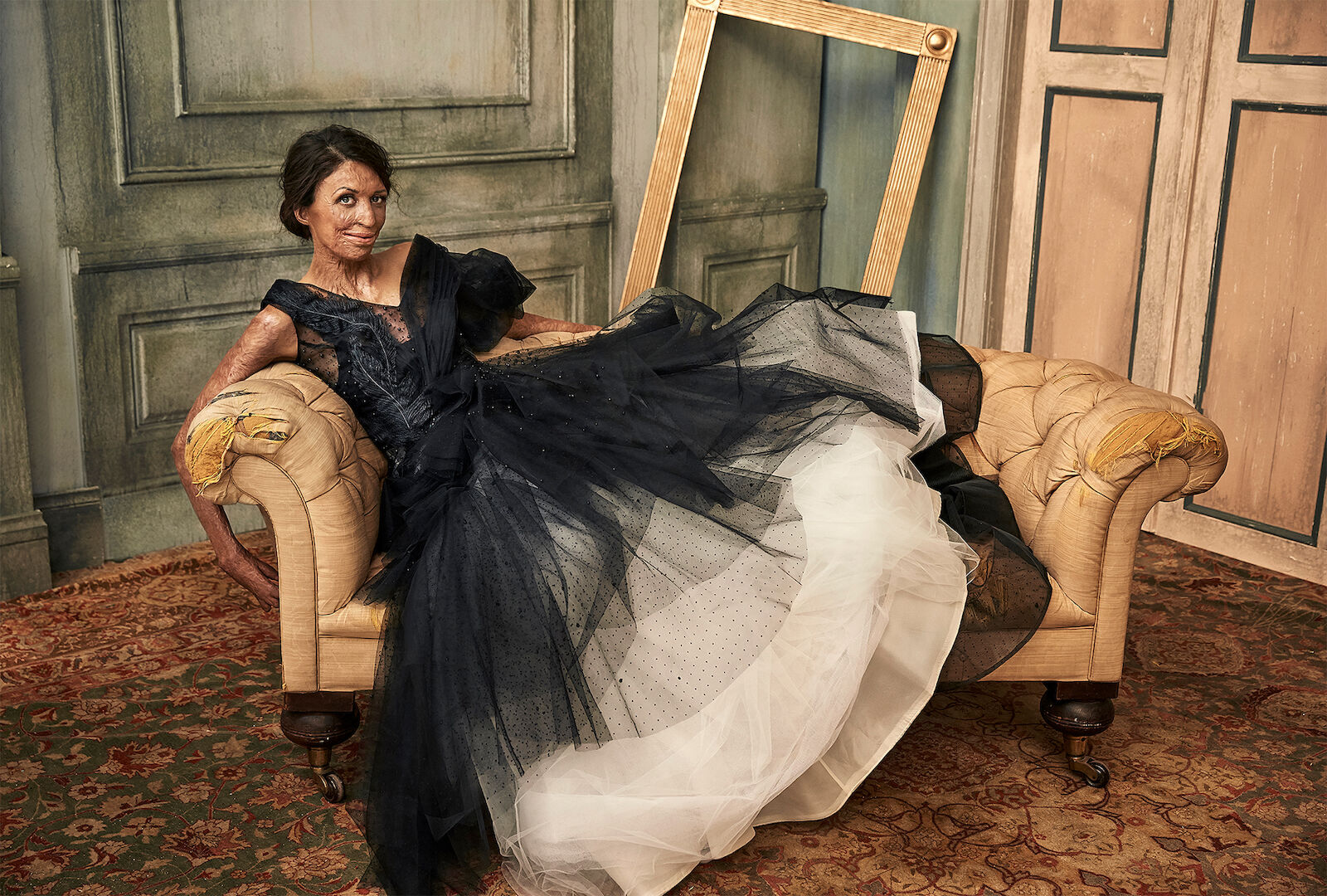 Elegant woman in a black and white gown reclines on a vintage couch next to an antique gold frame, indoors, for a fashion editorial.