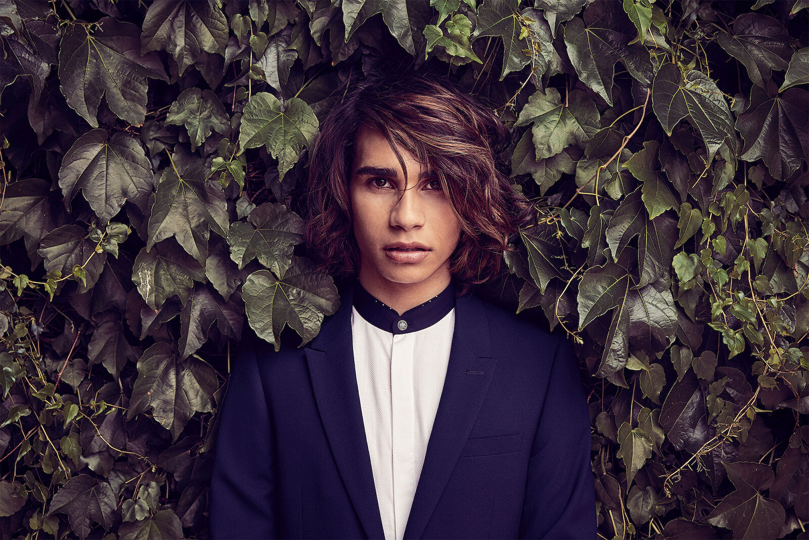 Headshot of a young man in a suit and white shirt posing against a dark green ivy wall in a portrait orientation with a serious expression.