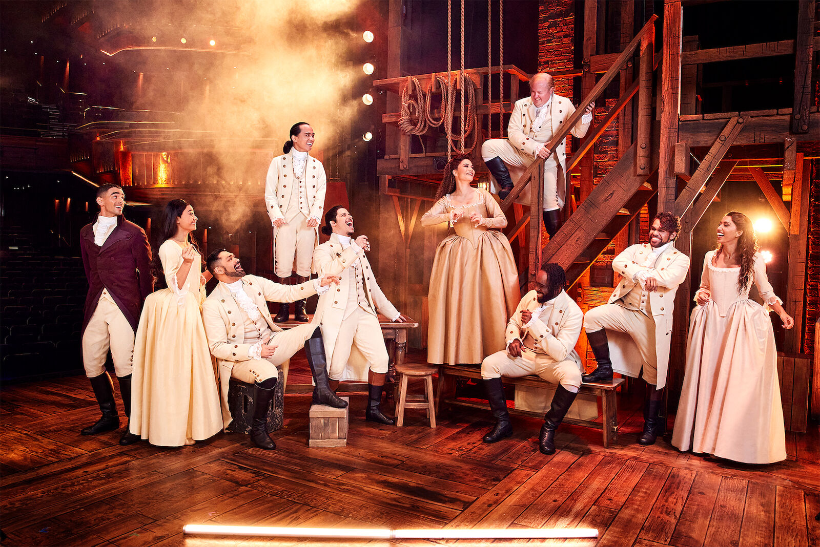 Group of actors onstage in historical costumes performing in the 'Hamilton' musical with wood stage and set, theatrical lighting.