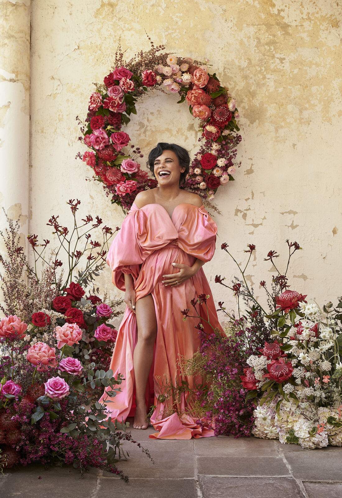 A happy pregnant woman poses in a pink gown against a textured wall, framed by a wreath and foreground of vibrant roses and greenery.