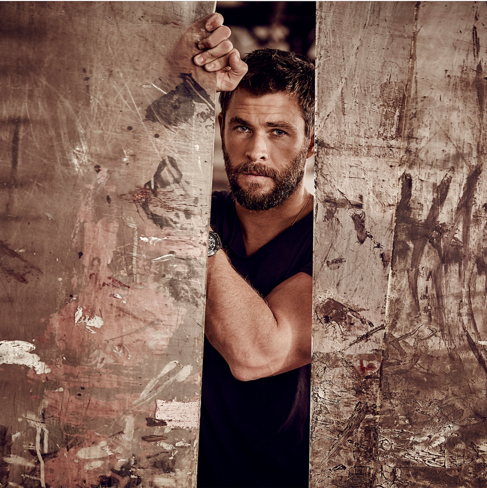 Close-up studio shot of Chris Hemsworth with a beard, wearing a black shirt, posing between two distressed boards.