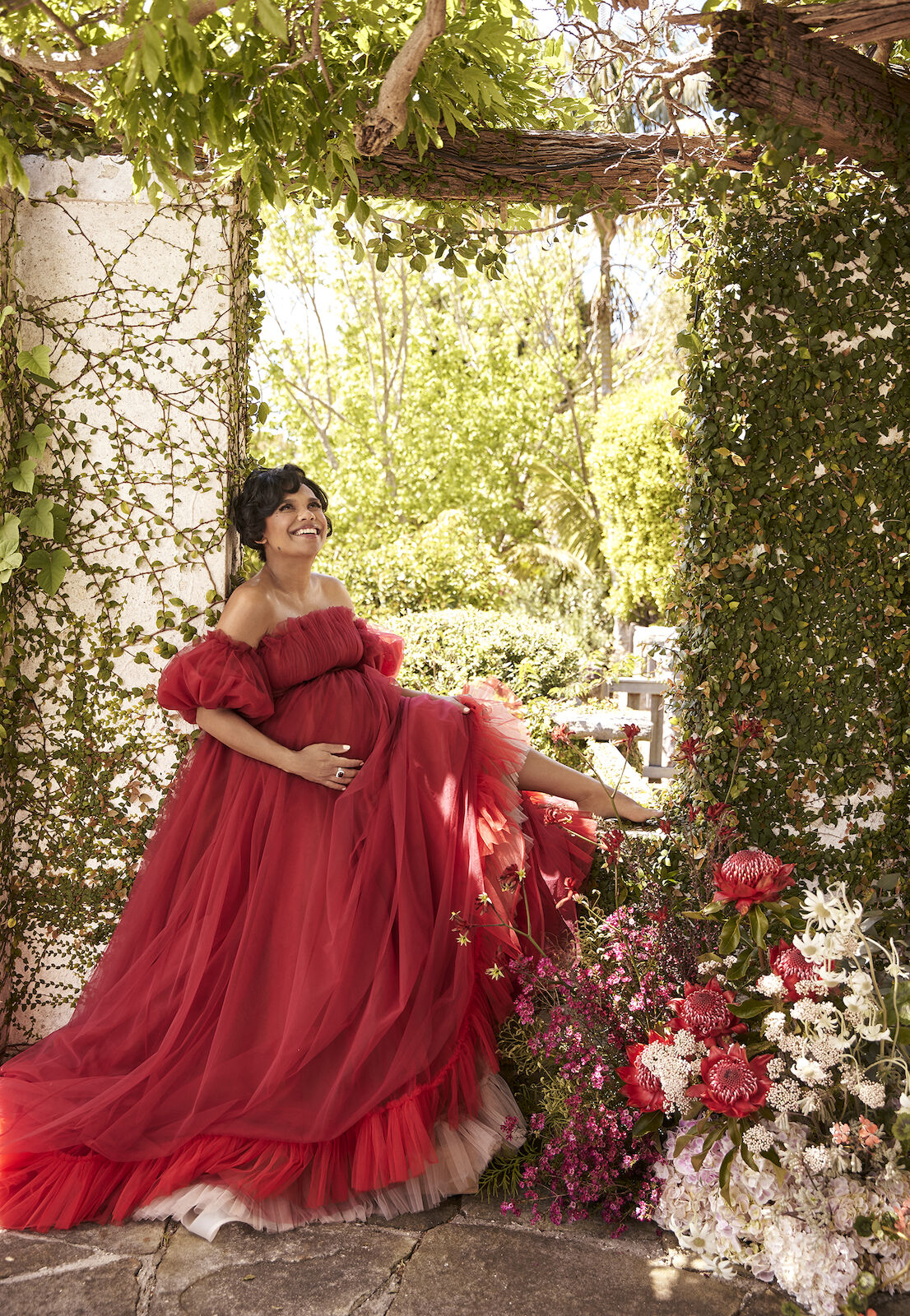 A pregnant woman in a red dress sits among flowers beneath a vine-covered trellis. The image highlights maternity, nature, and fashion.