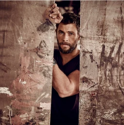 Close-up portrait of Chris Hemsworth peeking through a narrow opening between distressed wooden panels, photographed by Peter Brew Bevan.