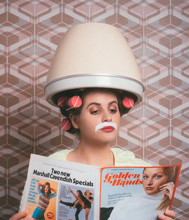 Woman with hair dryer and curlers reads a vintage fashion magazine with geometric patterned wallpaper in the background. Beauty concept.