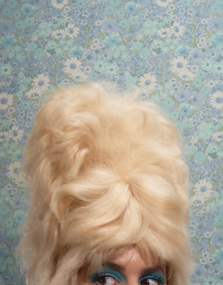 Glamorous woman with a bouffant hairstyle and bright teal eye makeup posing in front of a blue daisy floral background.