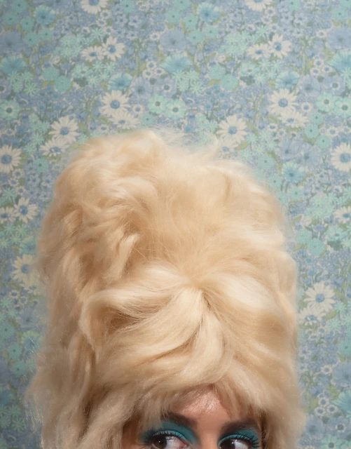 Glamorous woman with a bouffant hairstyle and bright teal eye makeup posing in front of a blue daisy floral background.
