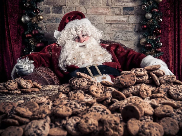 Santa Claus relaxes with a glass of milk, surrounded by piles of chocolate chip cookies in a cozy, festive, indoor setting.