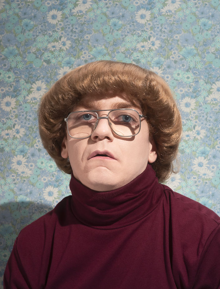 Headshot of a person wearing a wig, glasses, and a turtleneck sweater against a floral wallpaper backdrop in a portrait composition.