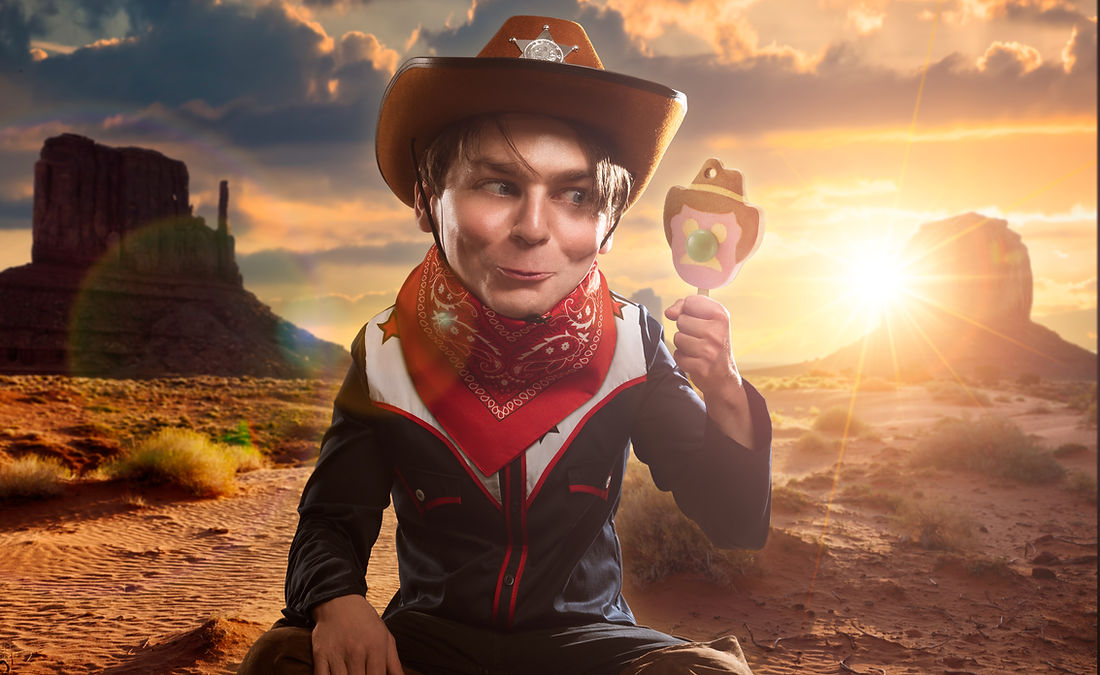 Young boy in cowboy costume holds a lollipop in a desert setting with rock formations and sunset; wearing hat, bandana, and vest.