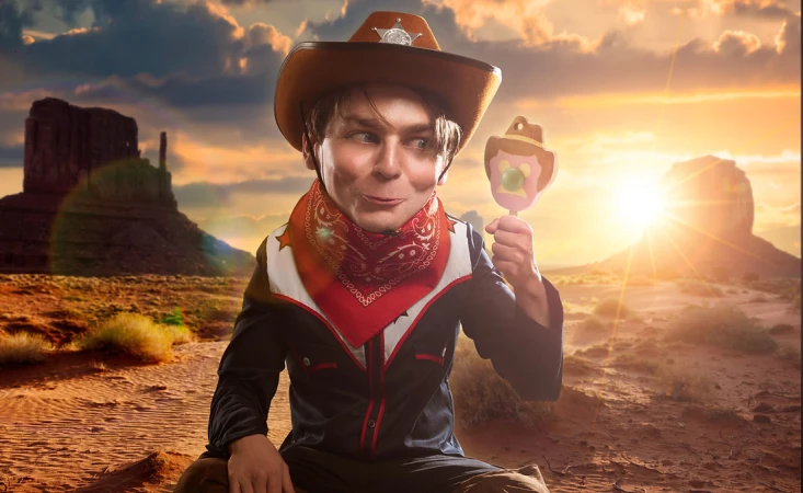 Young boy in cowboy costume holds a lollipop in a desert setting with rock formations and sunset; wearing hat, bandana, and vest.
