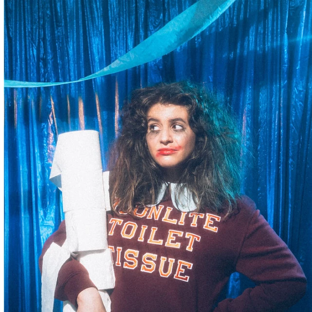 Adult woman in a maroon sweatshirt holds a stack of toilet paper and wears smeared red lipstick in front of a blue curtain.