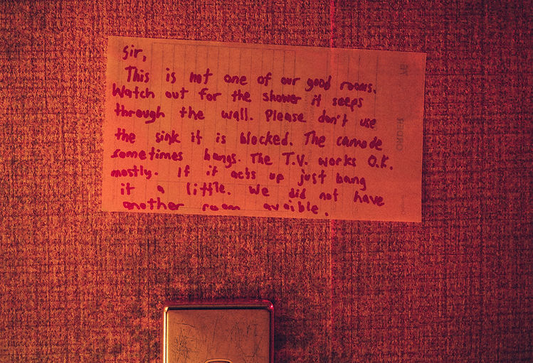 A handwritten note is taped to a textured, orange-toned wall, detailing issues with the room's shower, sink, and TV.