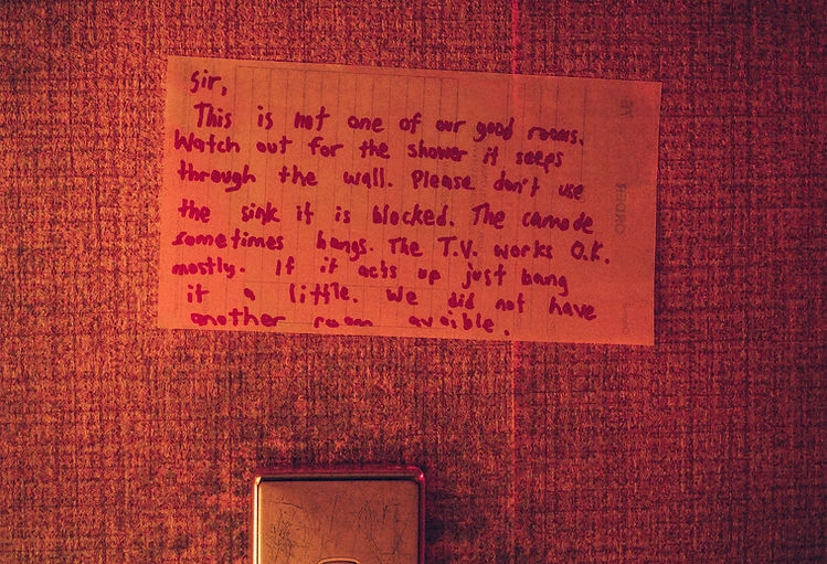 A handwritten note is taped to a textured, orange-toned wall, detailing issues with the room's shower, sink, and TV.