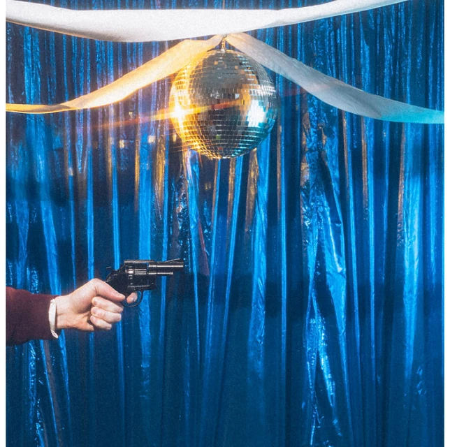 Hand with gun pointed at disco ball under white fabric, set against shiny blue draped background, creating a high-contrast, staged atmosphere.