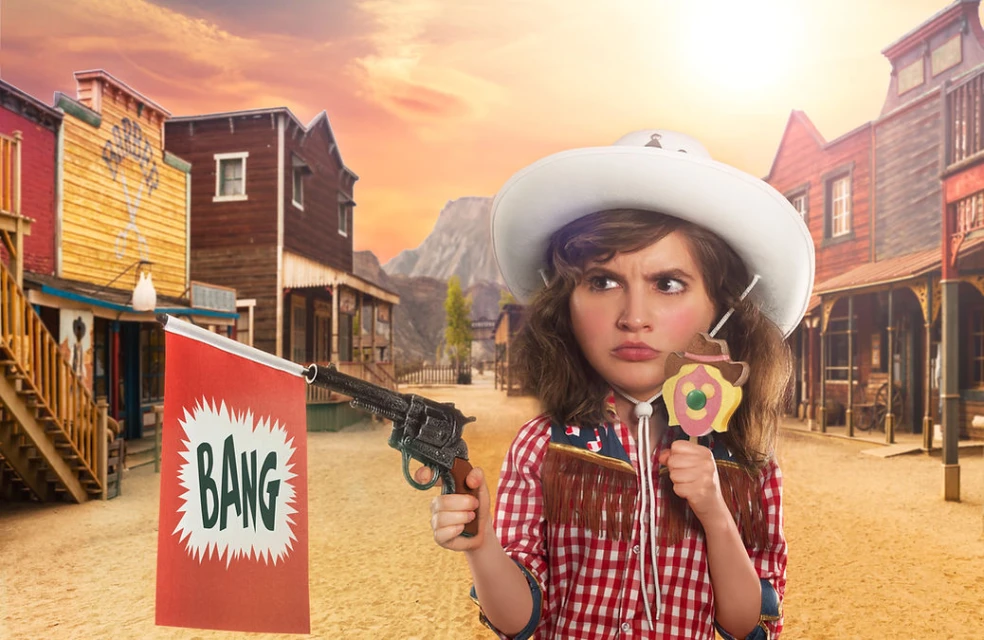 Young woman in a cowboy hat and red checkered shirt in a Western town holding a toy gun and lollipop, looking off to the side.