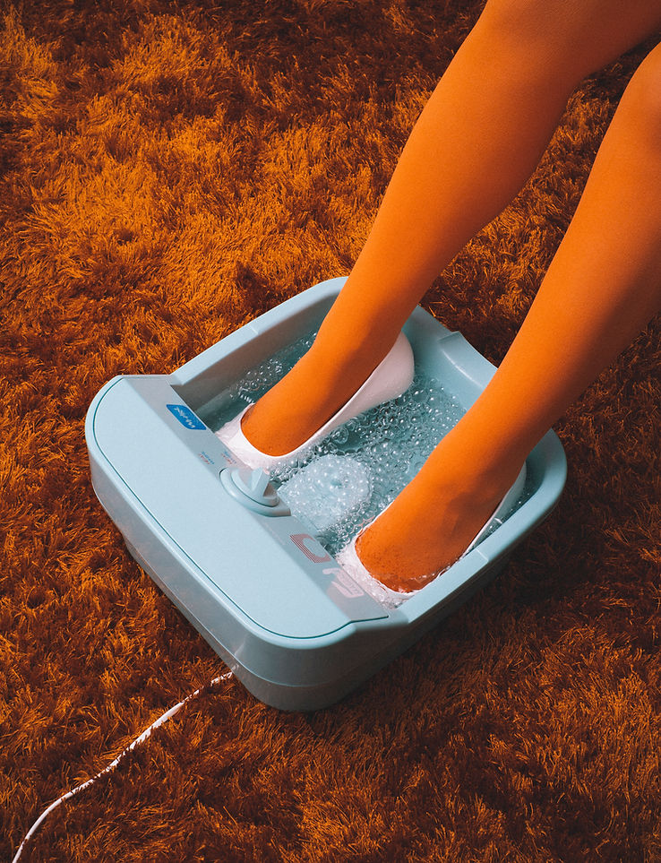 Woman soaks feet in a light blue water foot spa on top of an orange shag rug, showcasing relaxation and self-care.