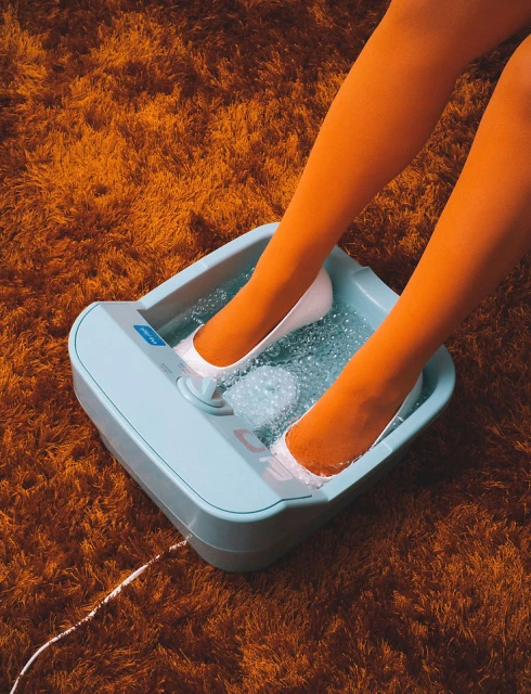 Woman soaks feet in a light blue water foot spa on top of an orange shag rug, showcasing relaxation and self-care.